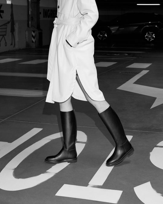 Black-and-white photo of a model walking in AERA ADA knee-high boots and a belted coat.