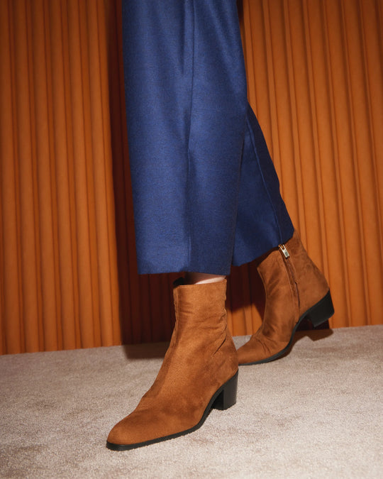 Brown suede ankle boots with block heel and side zipper, worn with blue pants on carpet
