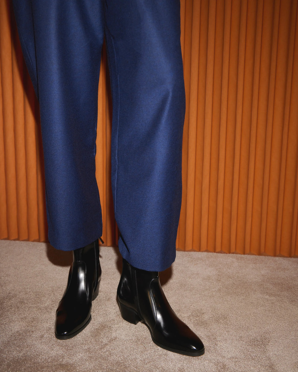 Shiny black leather ankle boots with blue tailored trousers on beige carpet, orange backdrop