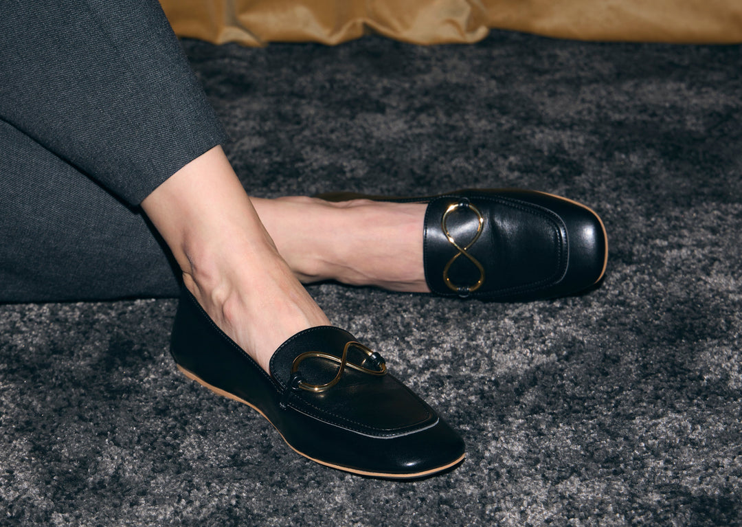 Mocassins noirs AERA ELLA ornés d’un motif infini doré, portés avec un pantalon anthracite sur un tapis foncé.