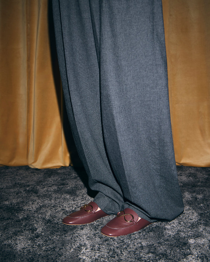 Modelo con mocasines castaños AERA ELLA y pantalón gris de pernera ancha, ante cortinas doradas.