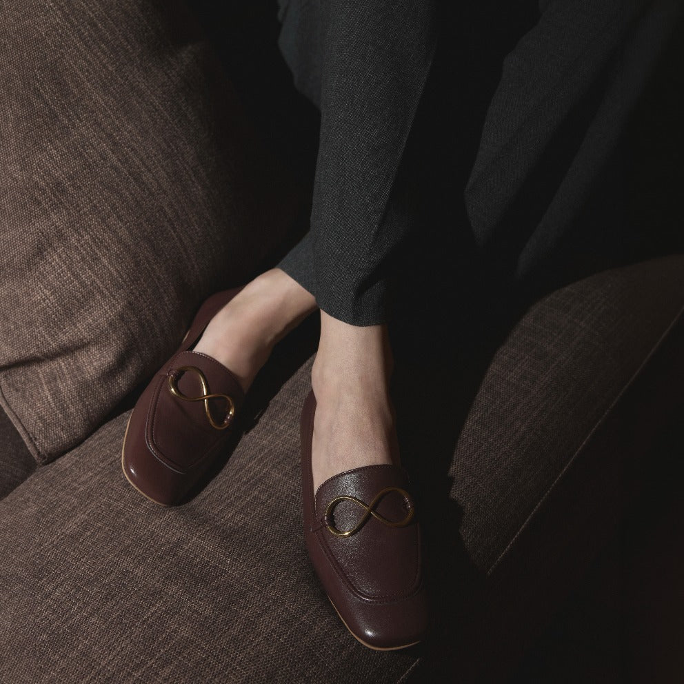 Mocasines castaños AERA ELLA con adorno dorado en forma de infinito, llevados por una modelo sentada en un sofá marrón.
