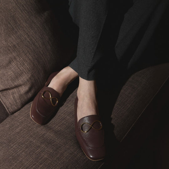 Mocasines castaños AERA ELLA con adorno dorado en forma de infinito, llevados por una modelo sentada en un sofá marrón.