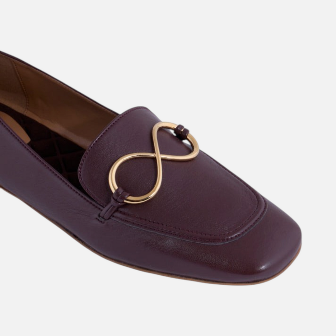 Mocasín AERA ELLA con acabado napa castaño, primer plano del adorno dorado en forma de infinito y la punta redondeada.