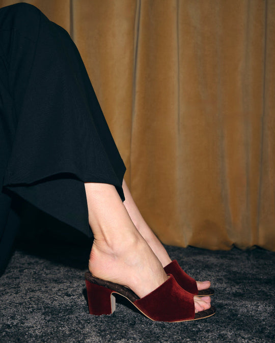 Model wearing AERA GIULIA cotto velvet block heel mules with black trousers on a dark carpet.